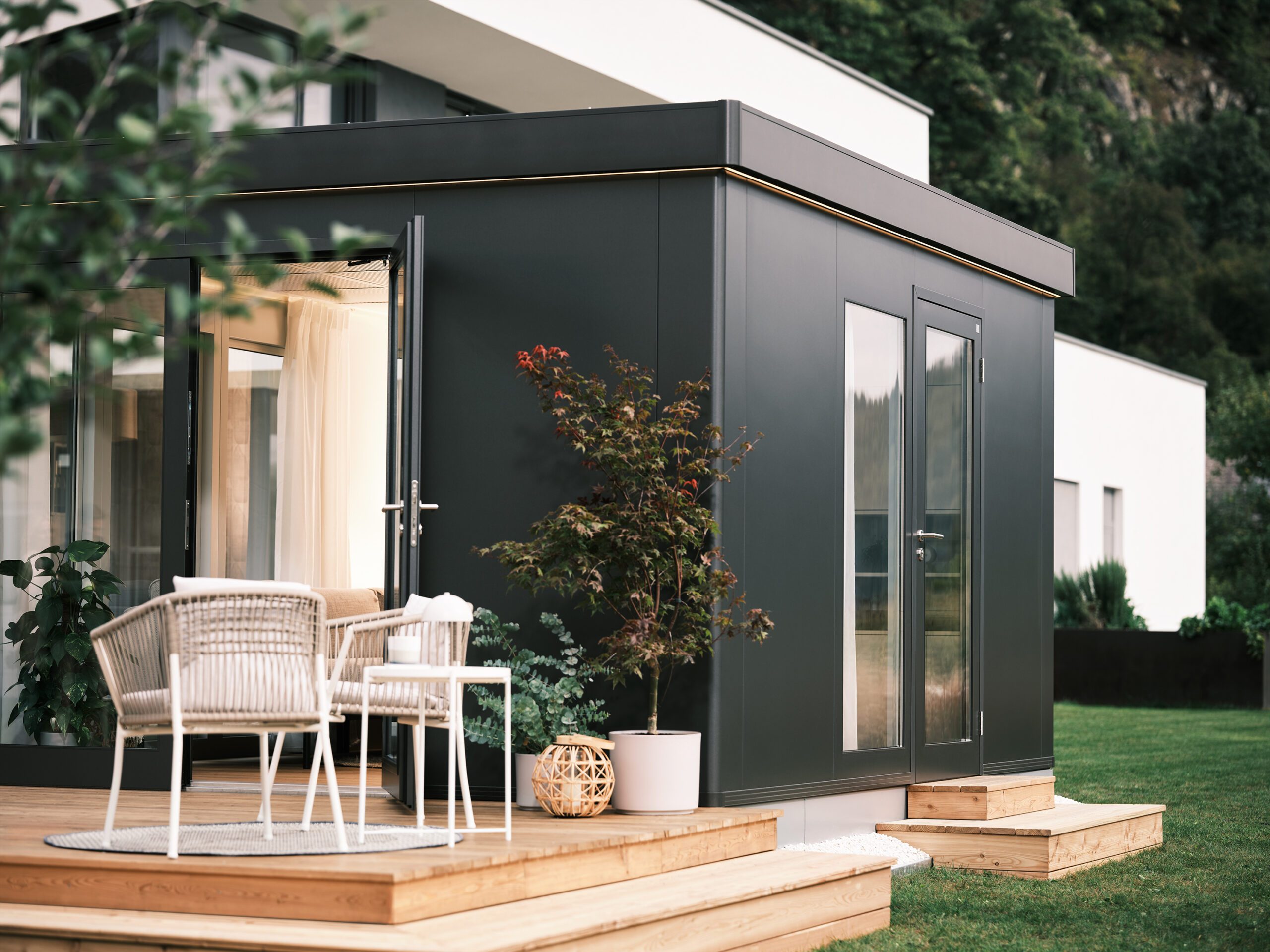 Seitenaufnahme eines dunkelgrauen Gartenhauses mit Flachdach, Ganzglastüren und Ganzglasfenster. Das Gartenhaus steht auf einem erhöhten Holzboden in einem Garten mit Gartensitzplatz. Erweiterter Wohnraum kaufen Schweiz.