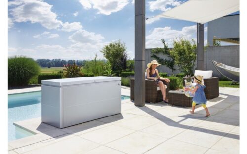 A grey metall box standing in front of a pool on a terrace. Next to the loungebox you can see a big garden lounge with a woman sitting on a chair and a child playing with its toys in front of her.